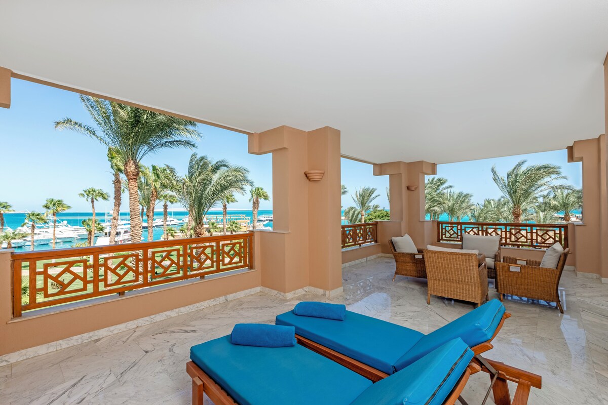 A balcony with blue lounge chairs, wicker furniture, and a view of the sea and palm trees.