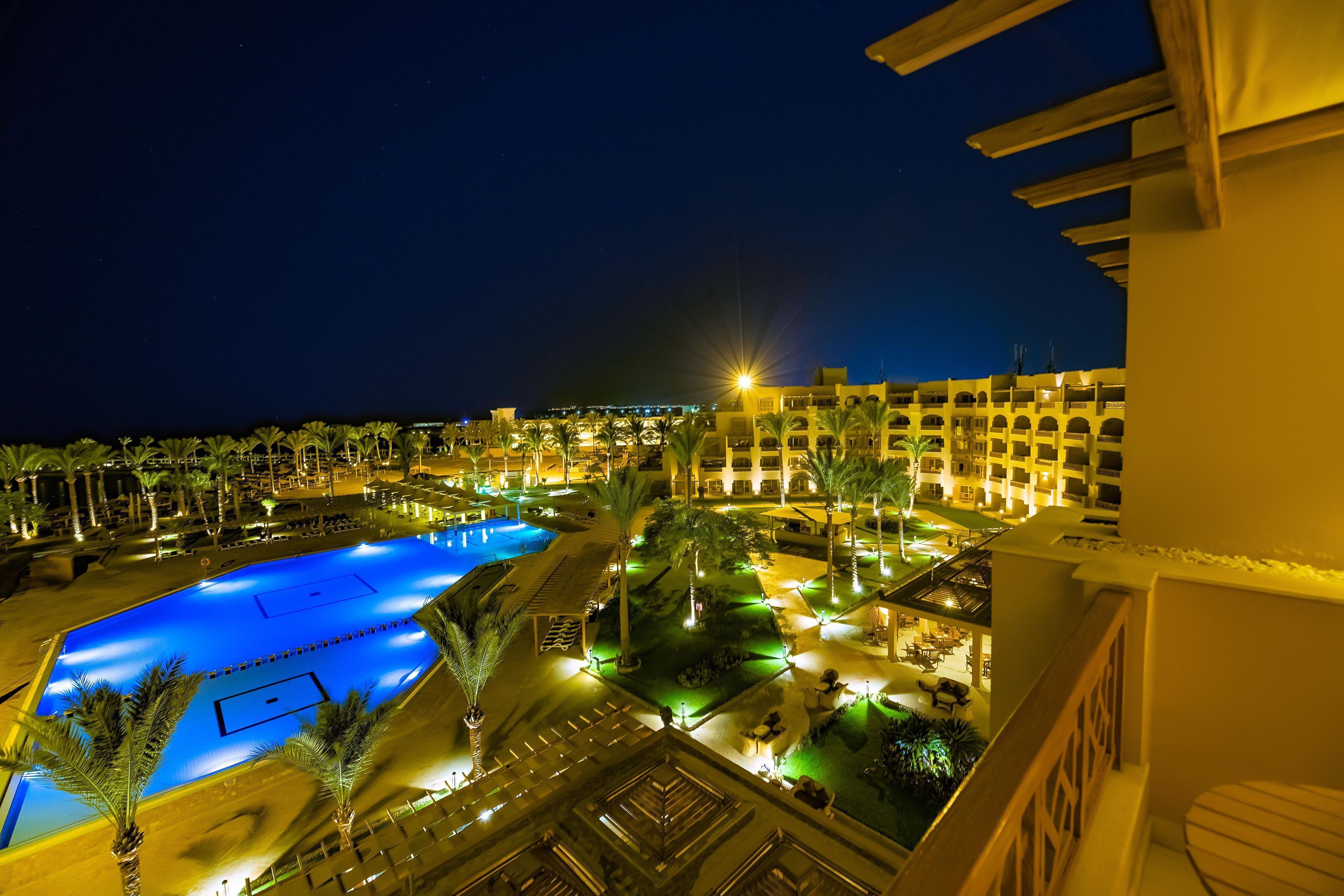 Vista nocturna de un resort con piscina, palmeras y edificios iluminados.