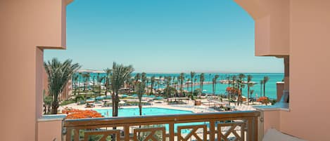 A balcony with wicker furniture, overlooking a pool and resort area with palm trees and a clear view of the sea.