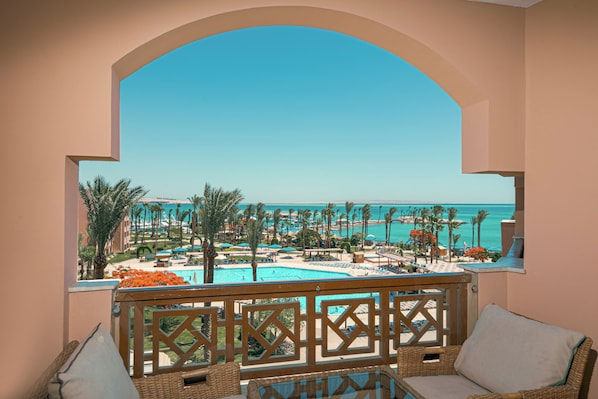 A balcony with wicker furniture, overlooking a pool and resort area with palm trees and a clear view of the sea.
