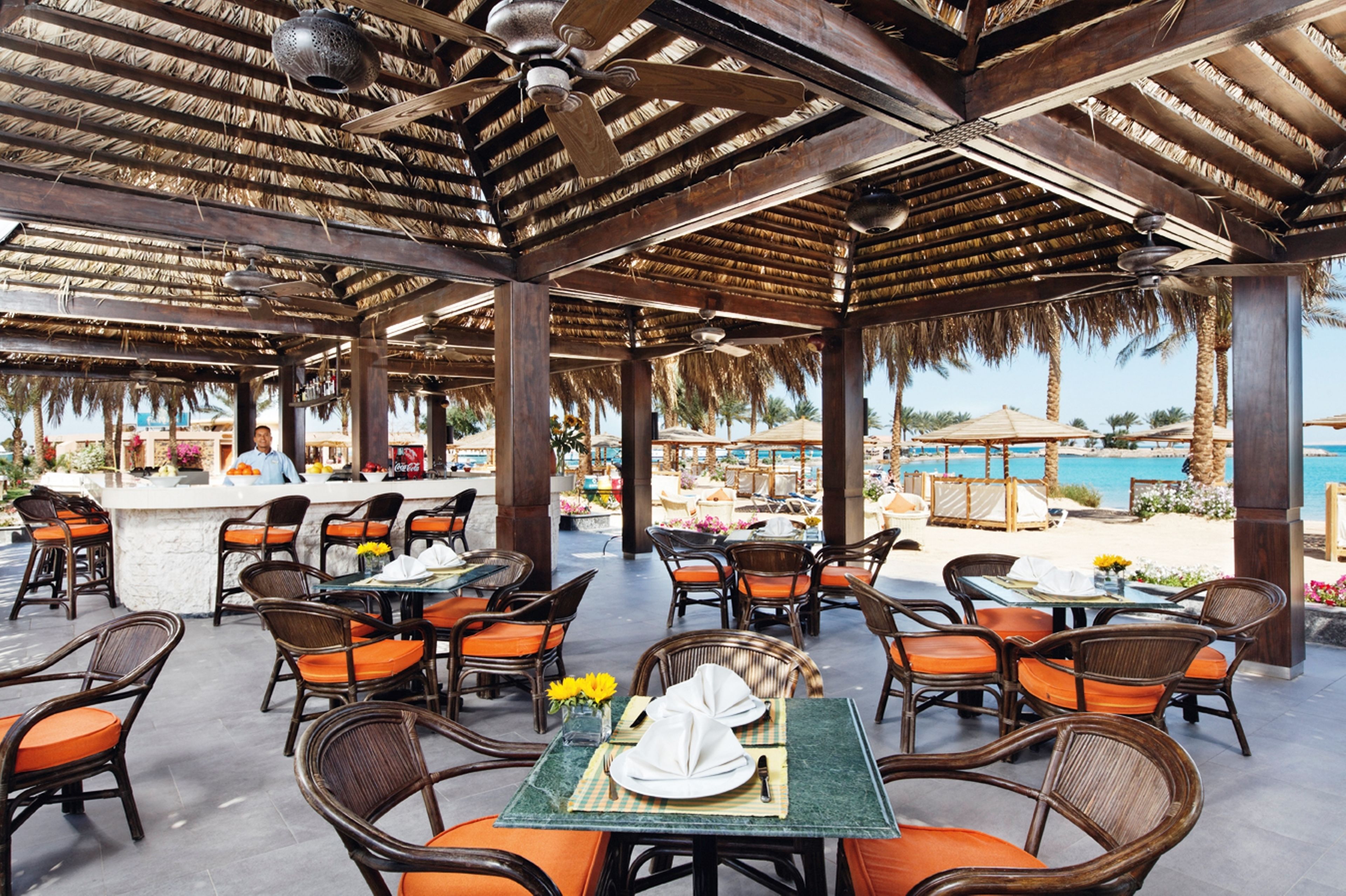 Ein Essbereich im Freien mit Korbstühlen und Tischen, eine Bar und Blick auf Strand und Meer.