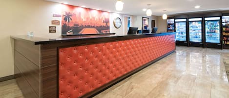Zona de recepción de un hotel con mostrador acolchado rojo, un mural con vista a la ciudad, un reloj y máquinas expendedoras.