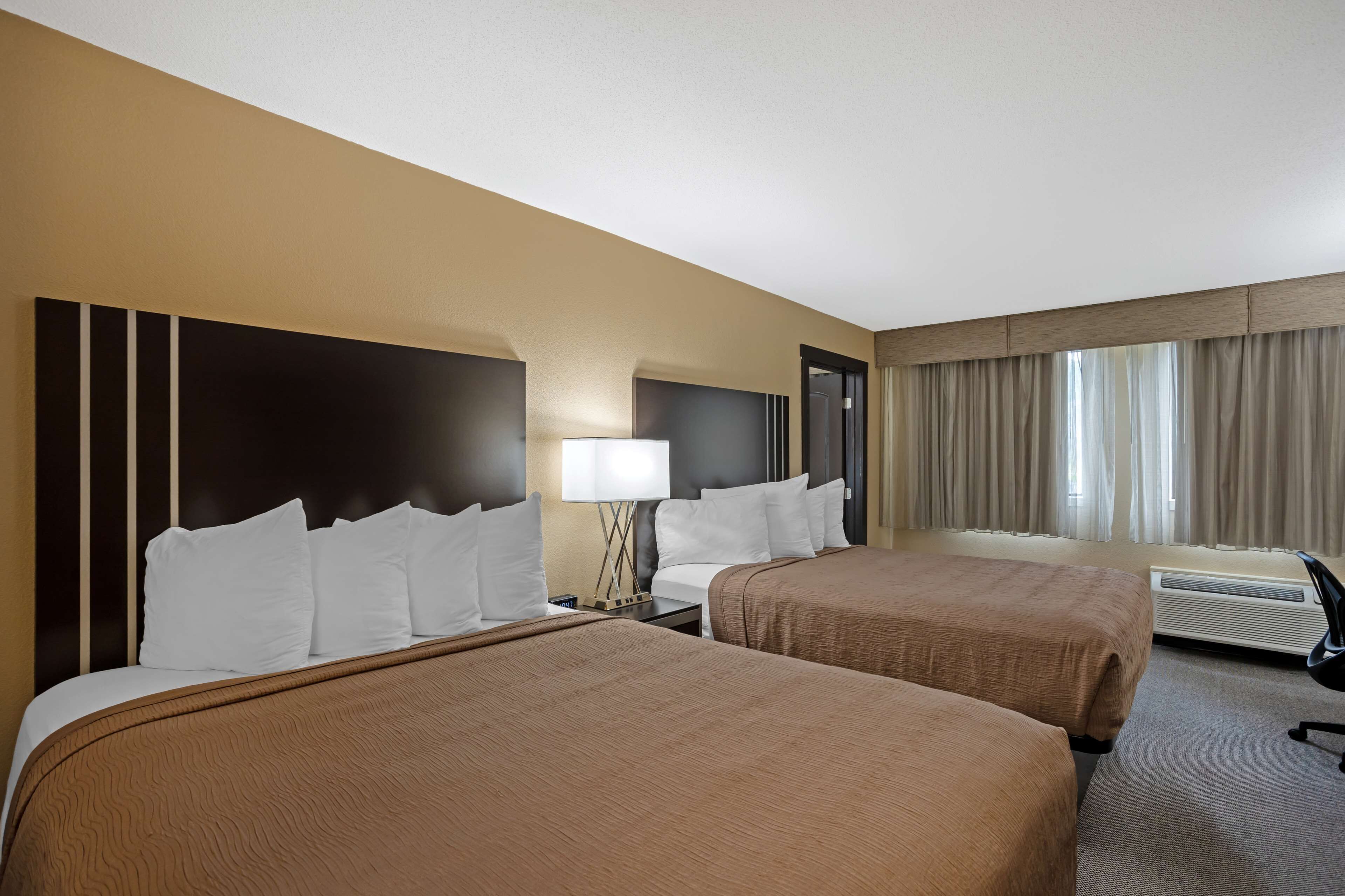 Habitación de hotel con dos camas, una mesita de noche, una lámpara, una silla y una ventana con cortinas.