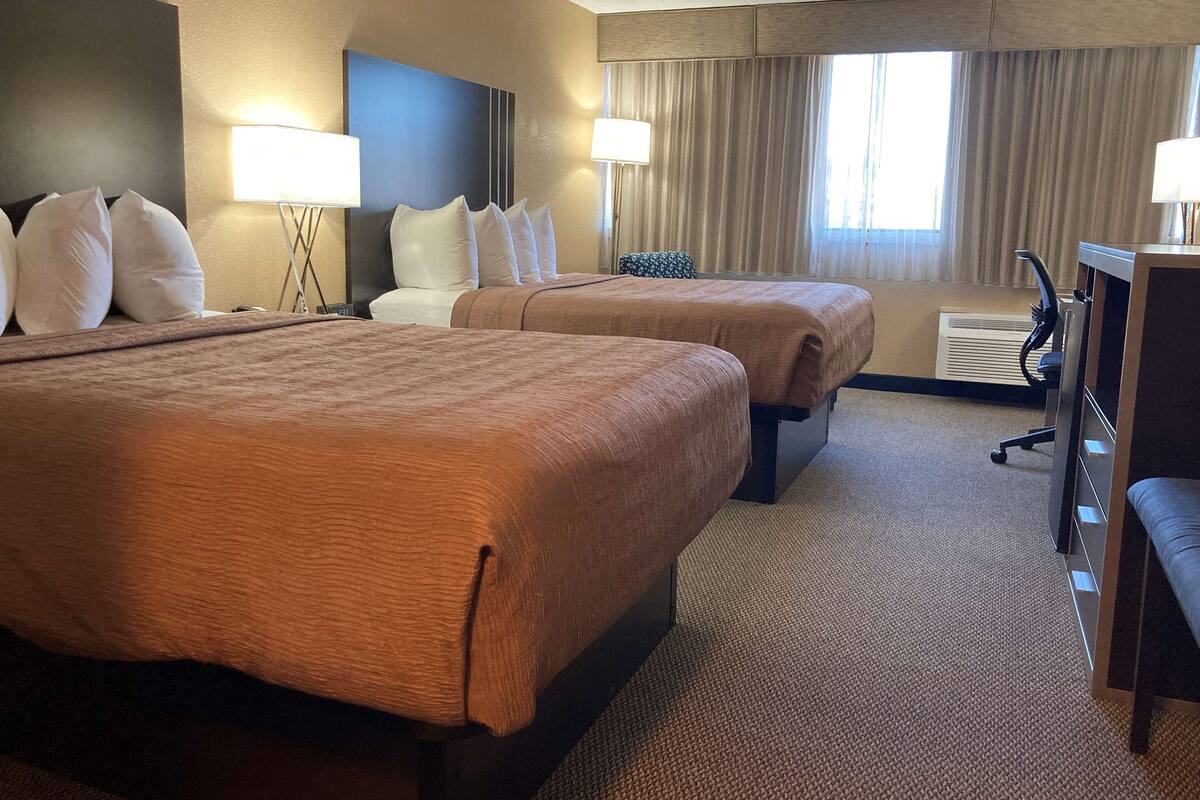 A hotel room with two beds, a desk, a chair, and a large window with curtains.