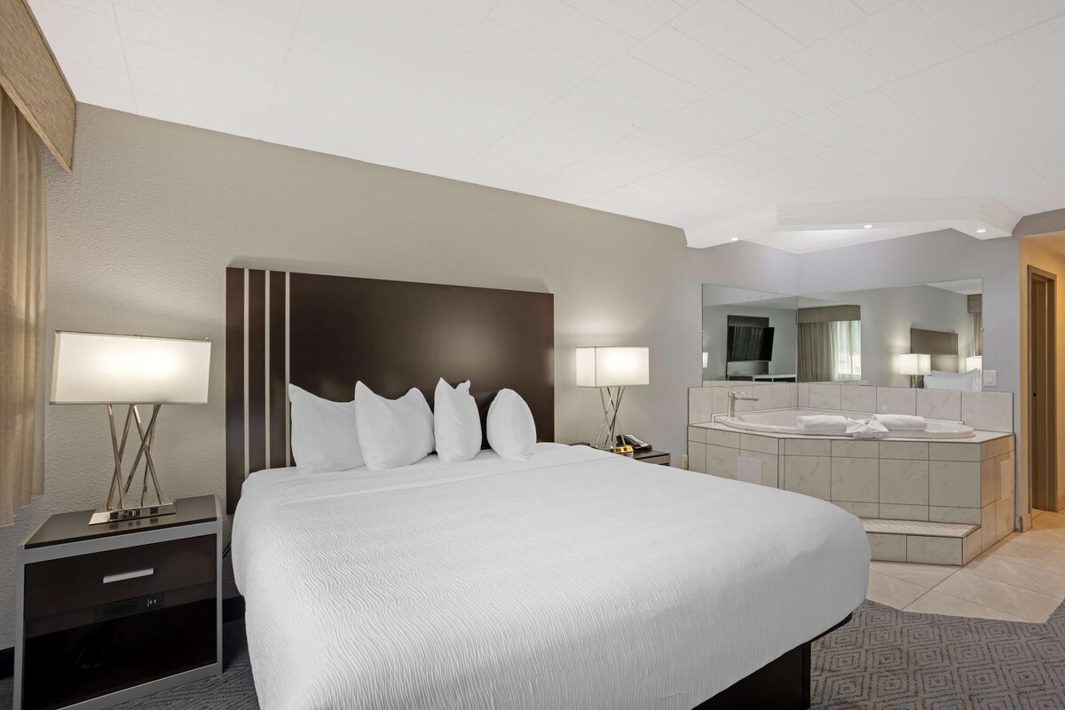 Habitación de hotel con una cama grande, lámparas de noche, una mesita de noche, un baño con bañera y un espejo grande.