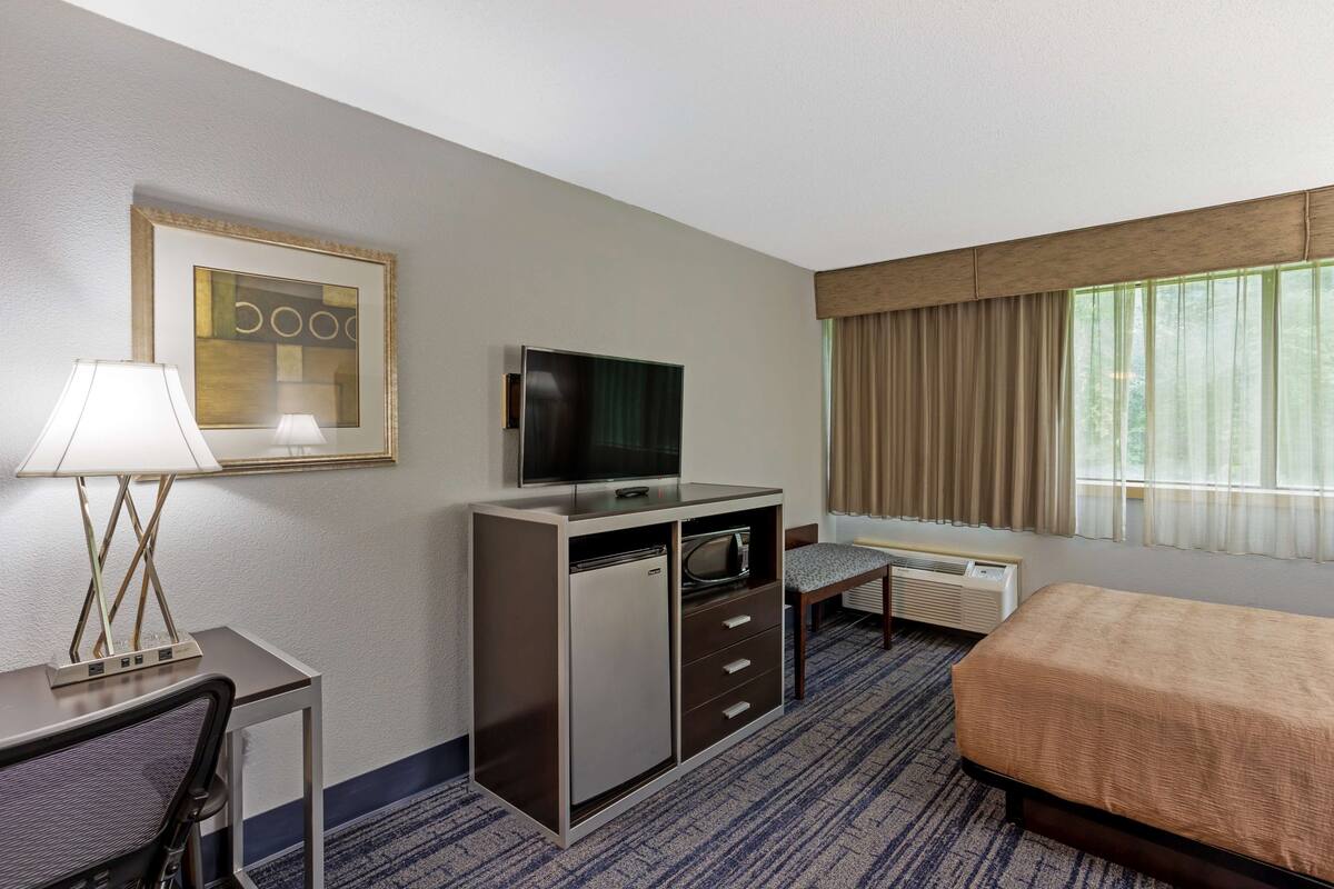 A hotel room with a bed, a TV, a desk with a lamp, a dresser, and a window with curtains.