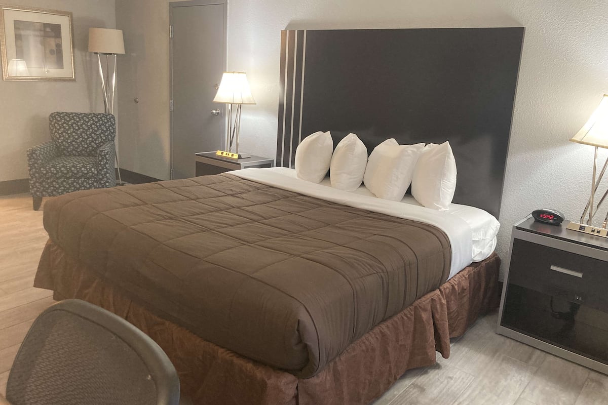 A hotel room with a large bed, a chair, a desk with a lamp, and a bedside table with a clock.