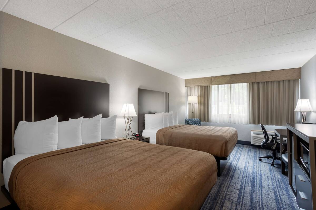 A hotel room with two beds, a desk, a chair, a lamp, and a window with curtains.