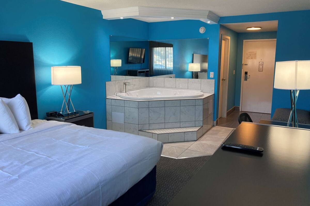 A hotel room with a large bed, a sitting area with a desk, a bathroom with a bathtub, and a TV mounted on the wall.
