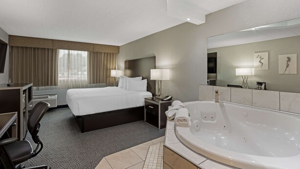 A hotel room with a large bed, a desk with a chair, a desk lamp, a bedside table with a phone and a lamp, a large bathtub, and a mirror on the wall. - Best Western Plus Reading Inn & Suites (Shillington)