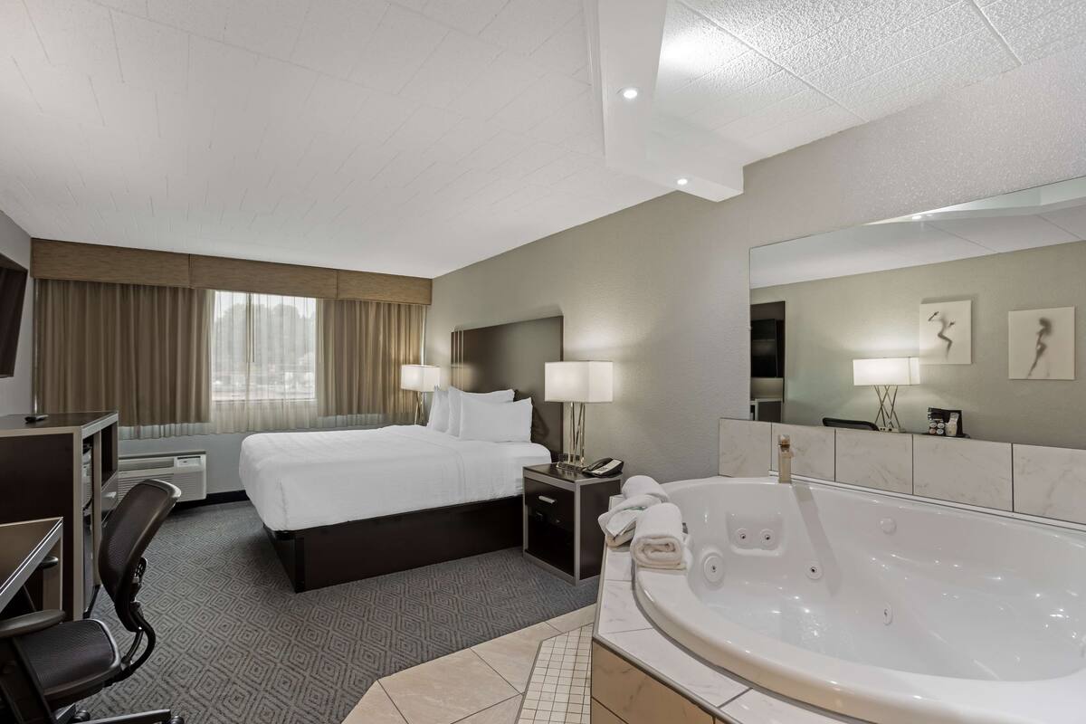 A hotel room with a large bed, a desk with a chair, a desk lamp, a bedside table with a phone and a lamp, a large bathtub, and a mirror on the wall.