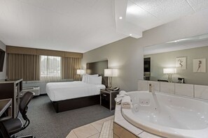 Ein Hotelzimmer mit einem großen Bett, einem Schreibtisch mit Stuhl, einer Schreibtischlampe, einem Nachttisch mit Telefon und Lampe, einer großzügigen Badewanne und einem Spiegel an der Wand.
