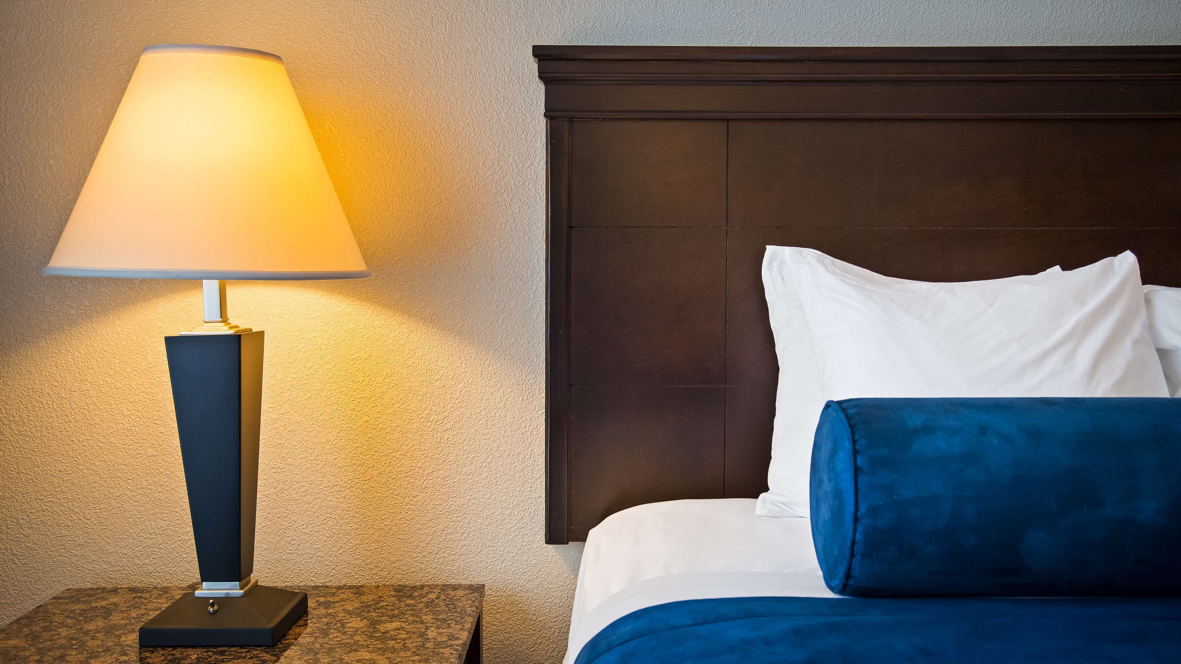 Una cama bien hecha con almohadones blancos y azules, un cabecero oscuro y una lámpara de mesa sobre una pared con textura.
