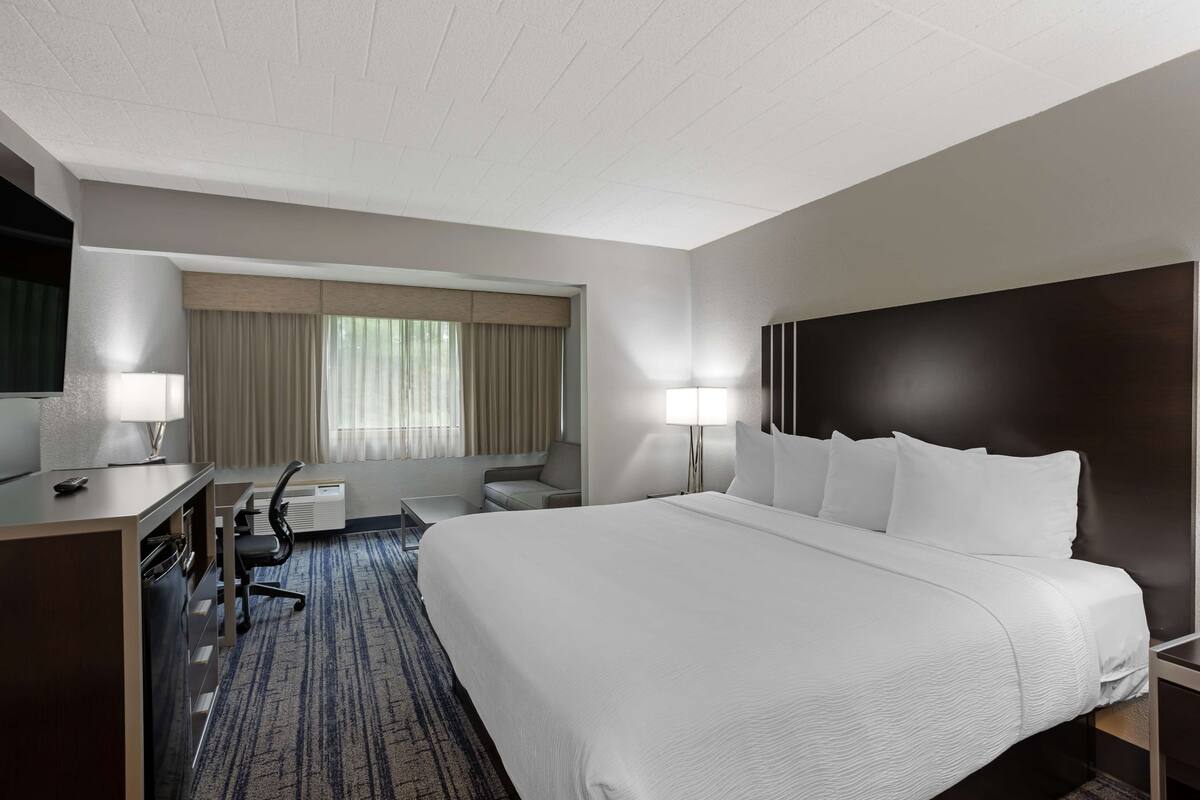 A hotel room with a large bed, a desk with a chair, a lamp, and a window with curtains.