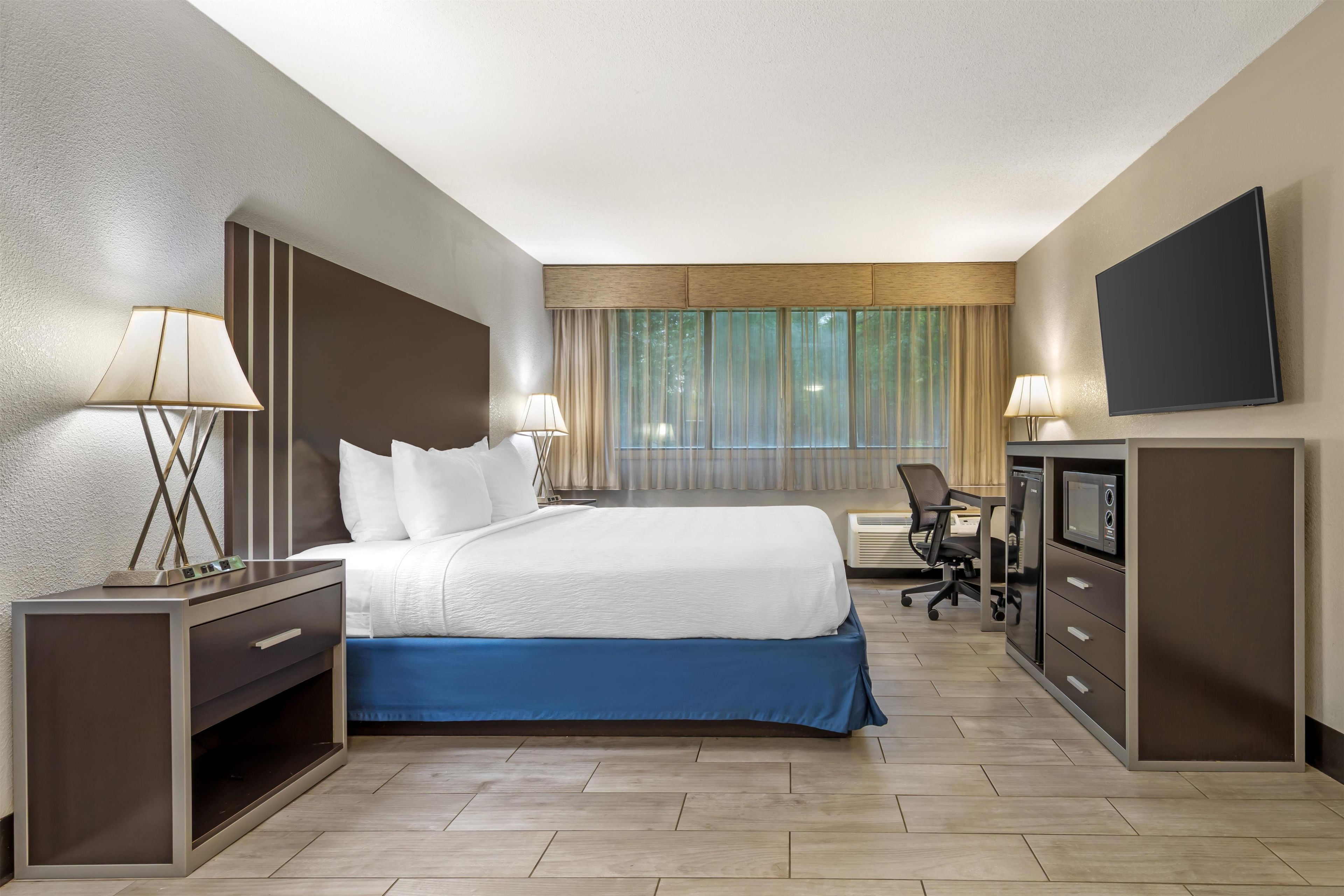 A hotel room with a bed, bedside tables, a desk, a chair, a television, and a window with curtains.