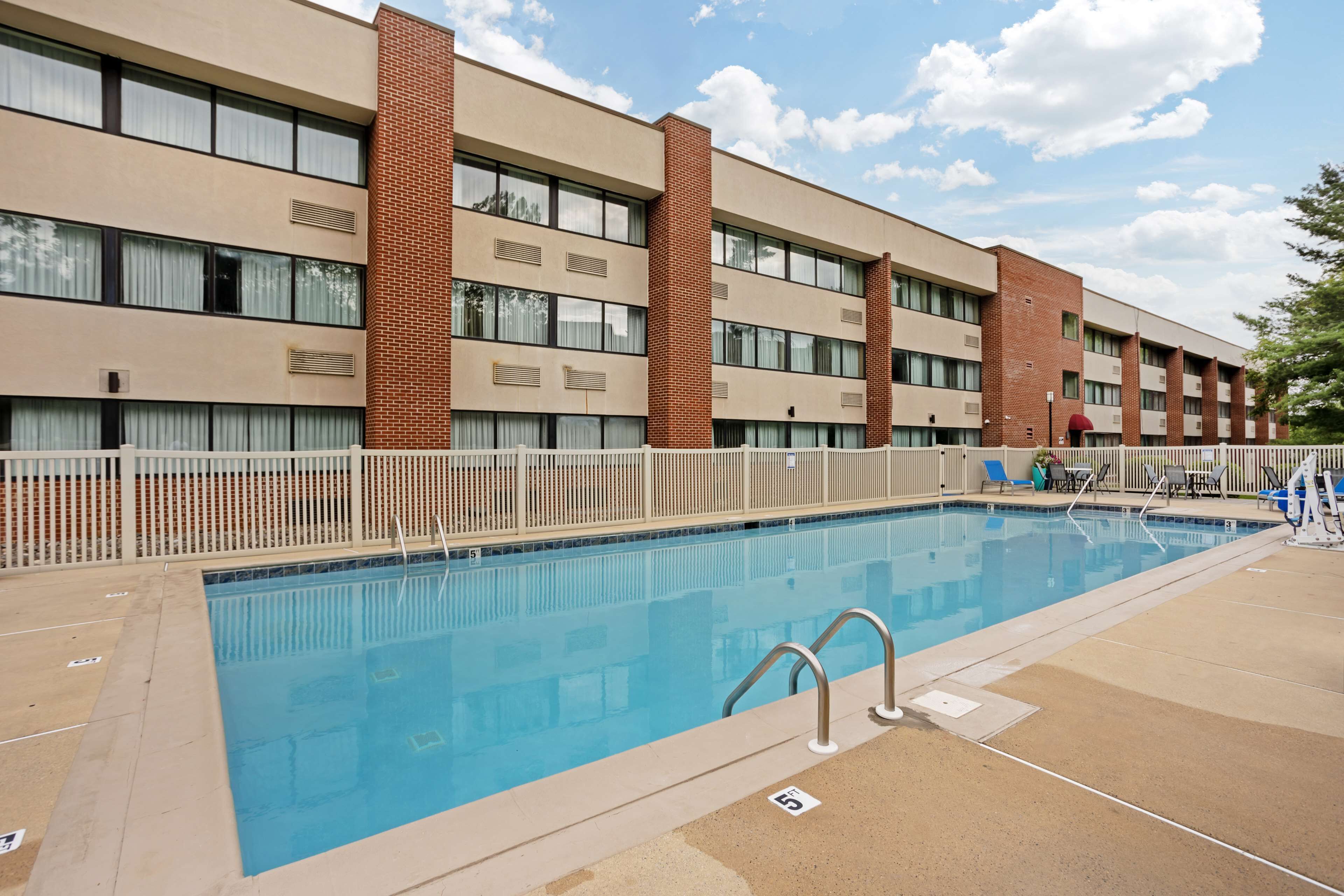Una piscina rectangular con valla blanca, rodeada por un edificio con múltiples ventanas y pilares de ladrillo.