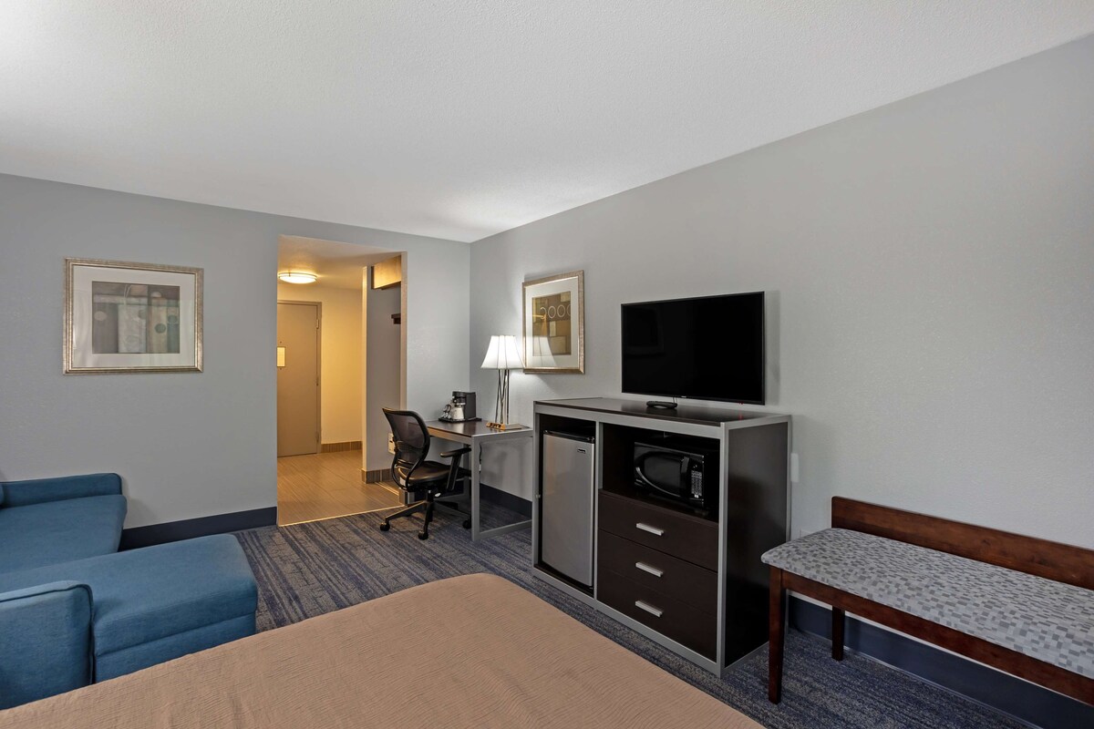 Habitación de hotel con cama, escritorio con computadora, televisor, silla y refrigerador pequeño.