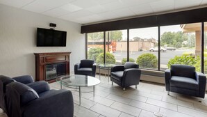 A modern lounge area with a fireplace, comfortable seating, a flat-screen TV, and large windows offering a view of the parking lot and buildings outside. - Best Western Plus Reading Inn & Suites (Shillington)