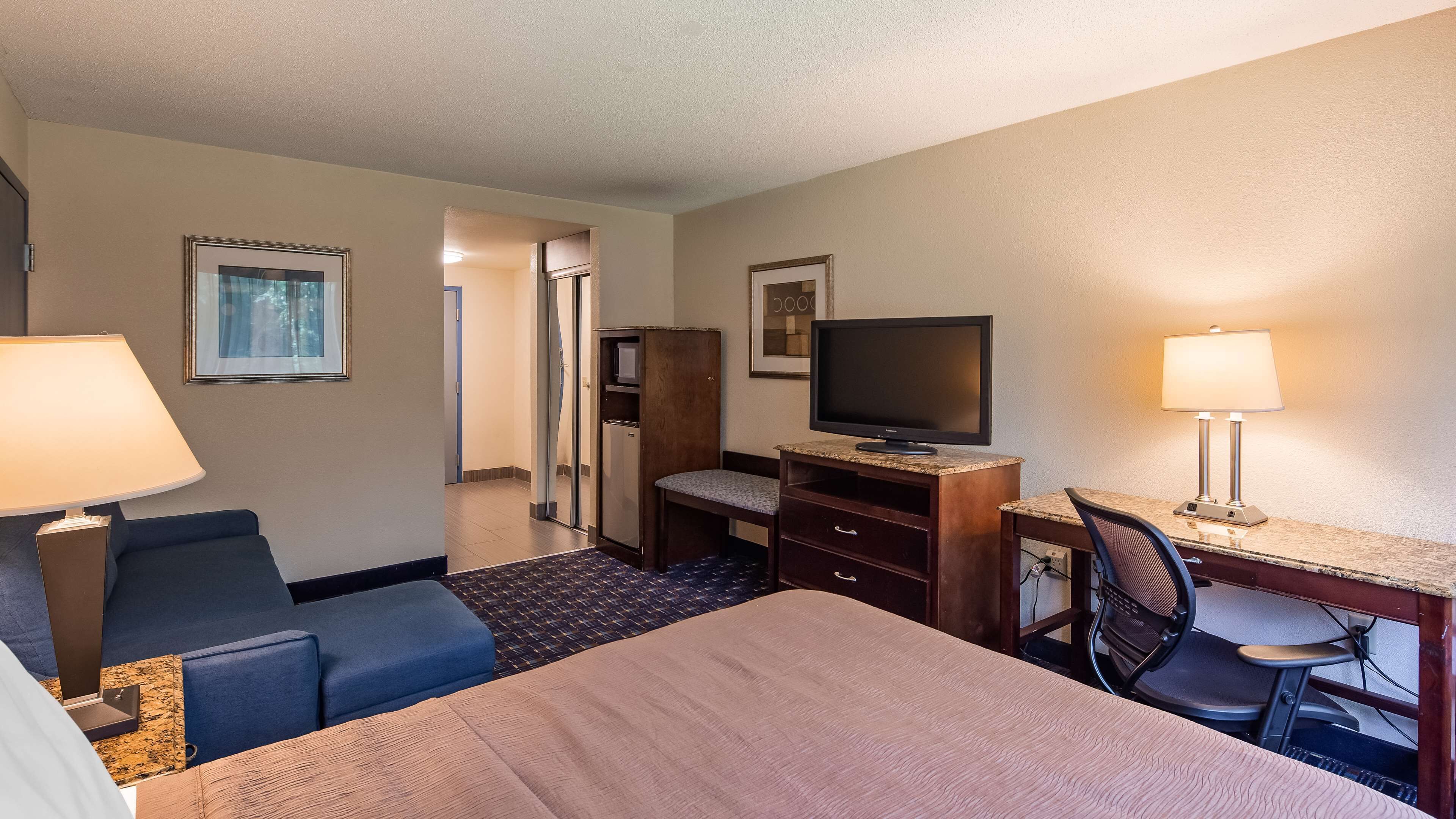 A hotel room with a bed, a desk with a computer, a television, a chair, a desk lamp, and a small cabinet.