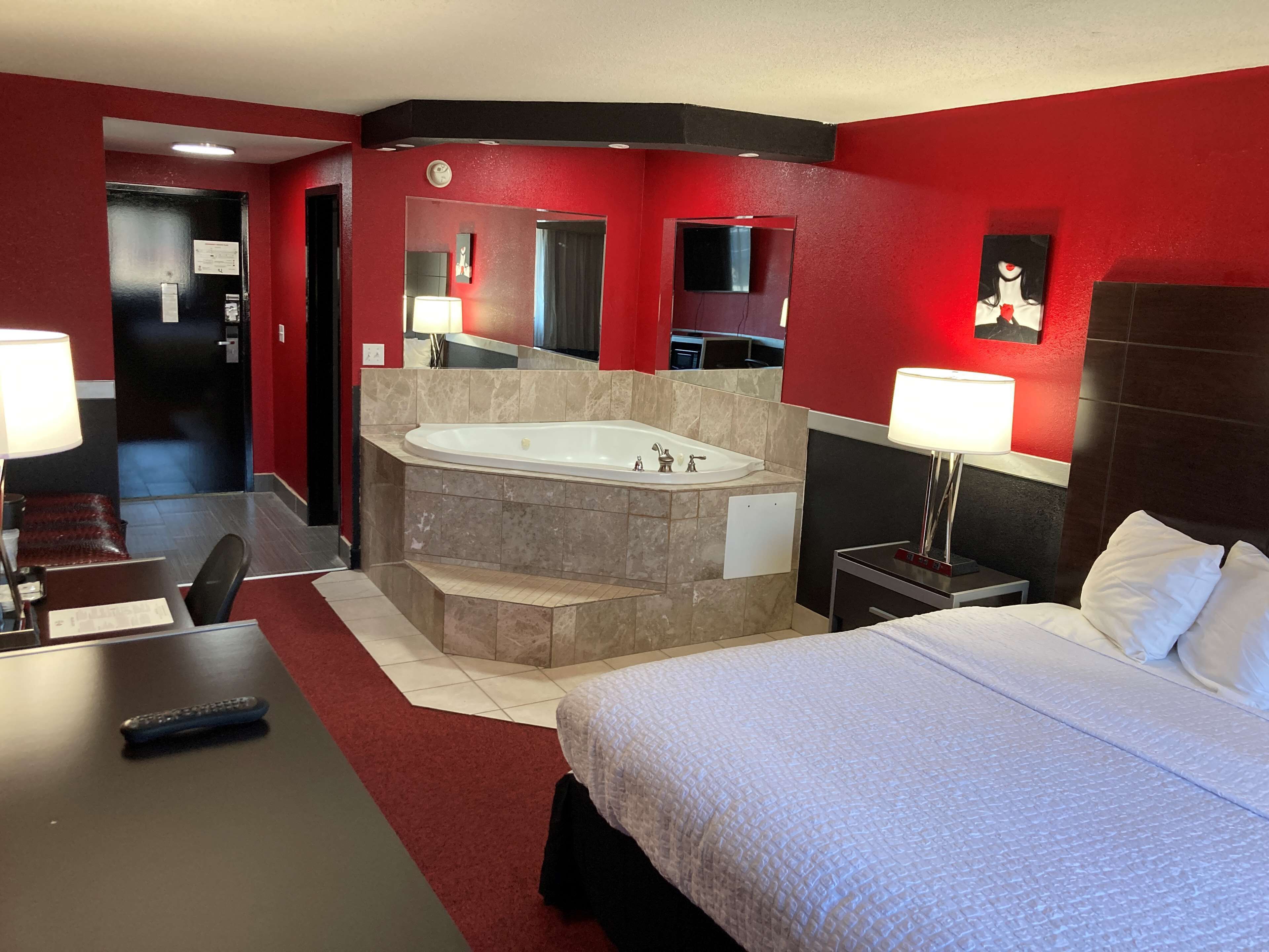 A hotel room with a large bed, a separate bathroom with a bathtub, a desk with a chair, and a small table with a lamp.