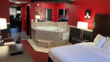 A hotel room with a large bed, a separate bathroom with a bathtub, a desk with a chair, and a small table with a lamp.