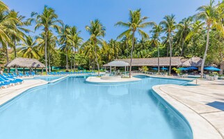 Una piscina grande con sillas reclinables azules y estructuras de techo de paja.