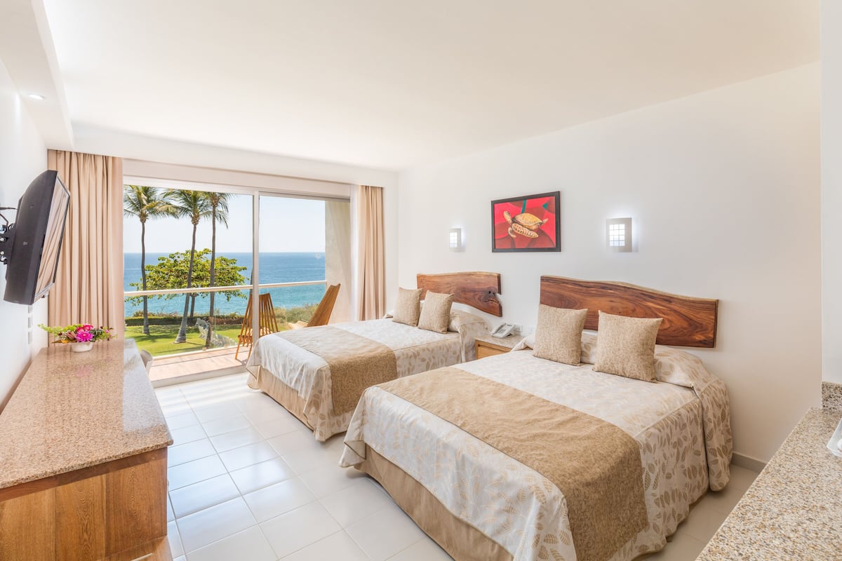 A hotel room with two beds, a TV, a desk, and a balcony with a view of the ocean.
