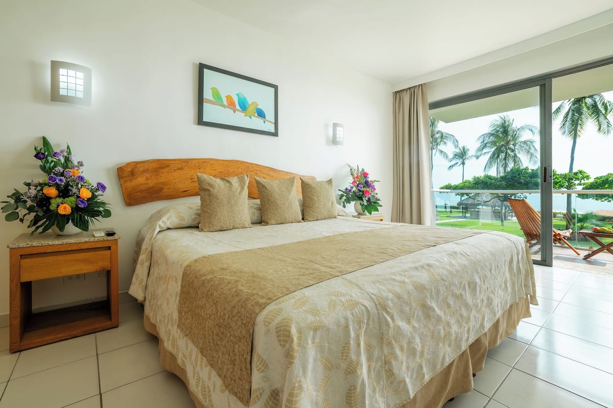 A hotel room with a large bed, a wooden headboard, a nightstand with a vase of flowers, a painting of birds, and a view of a tropical landscape through a sliding glass door.