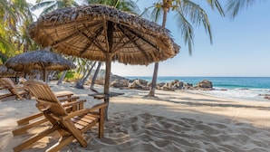 A beach with wooden lounge chairs and thatched umbrellas, a clear sky, and a rocky shoreline. - Posada Real Puerto Escondido (Puerto Escondido)