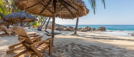 Una playa con sillones de madera y sombrillas de paja, cielo despejado y un litoral rocoso.