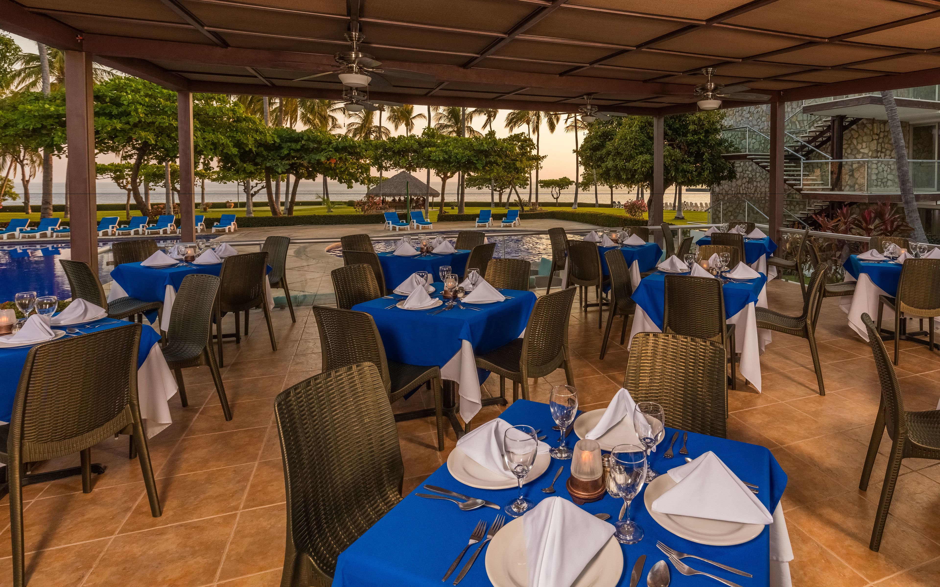 An outdoor dining area with tables set for two, covered by a roof, and overlooking a pool and palm trees.