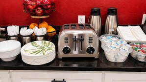 A breakfast buffet setup with a toaster, fruit bowl, coffee makers, and stacked plates. - Econo Lodge Cranston - Providence (Cranston)
