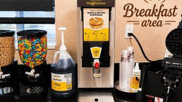 Una zona de desayuno con un dispensador de masa para gofres, dispensadores de cereales y botellas de jarabe para gofres.