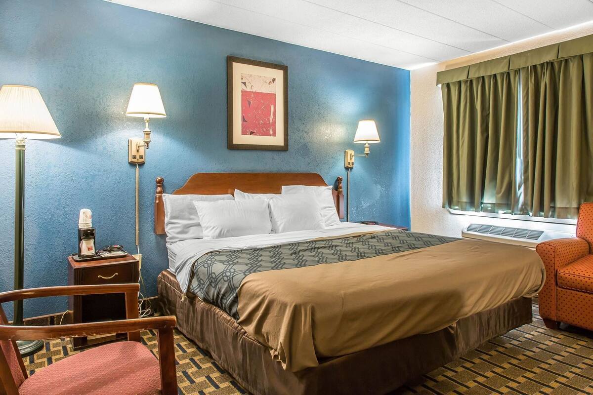 A hotel room with a bed, two bedside lamps, a framed artwork on the wall, a window with curtains, a chair, and a small table with a lamp.