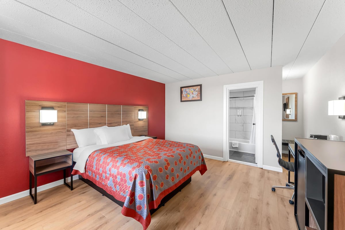 A modern bedroom with a red accent wall, a wooden headboard, a bed with a patterned bedspread, a nightstand, a desk with a chair, and a bathroom visible through an open door.
