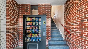 A vending machine with snacks and a small window, located next to a staircase in a building with brick walls. - Econo Lodge Cranston - Providence (Cranston)