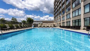 Una piscina con sillones de descanso y sombrillas, rodeada por un edificio alto con numerosas ventanas.