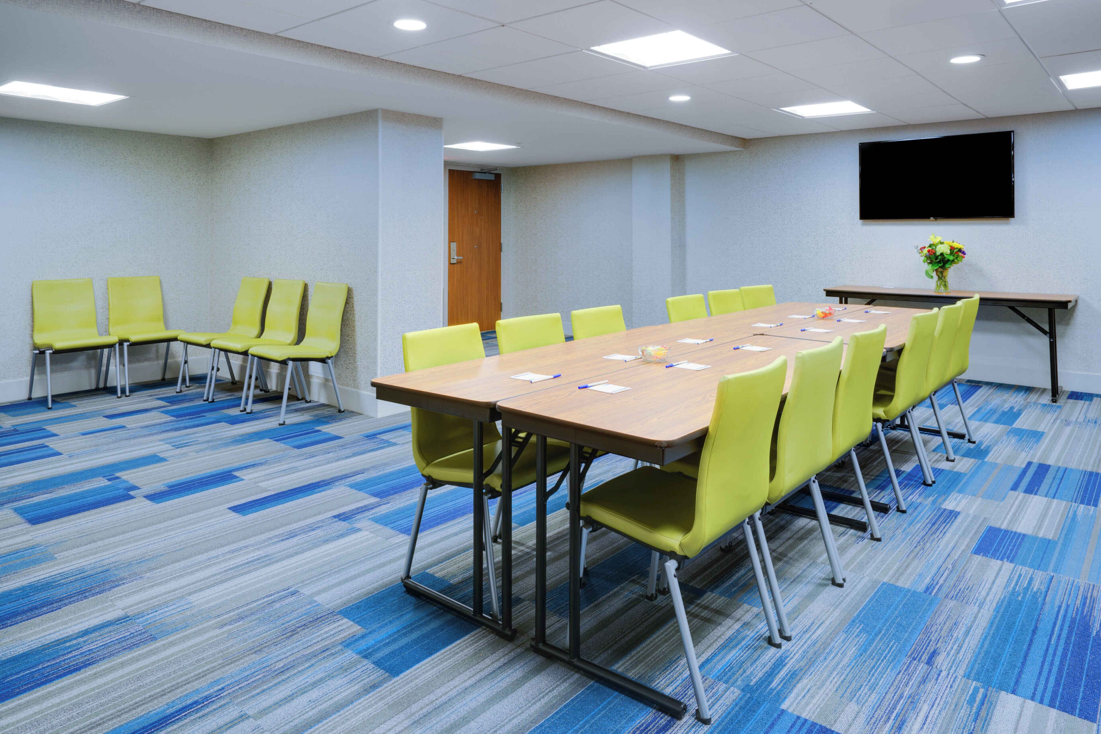 A conference room with a long table, multiple chairs, a projector screen, and a carpeted floor.