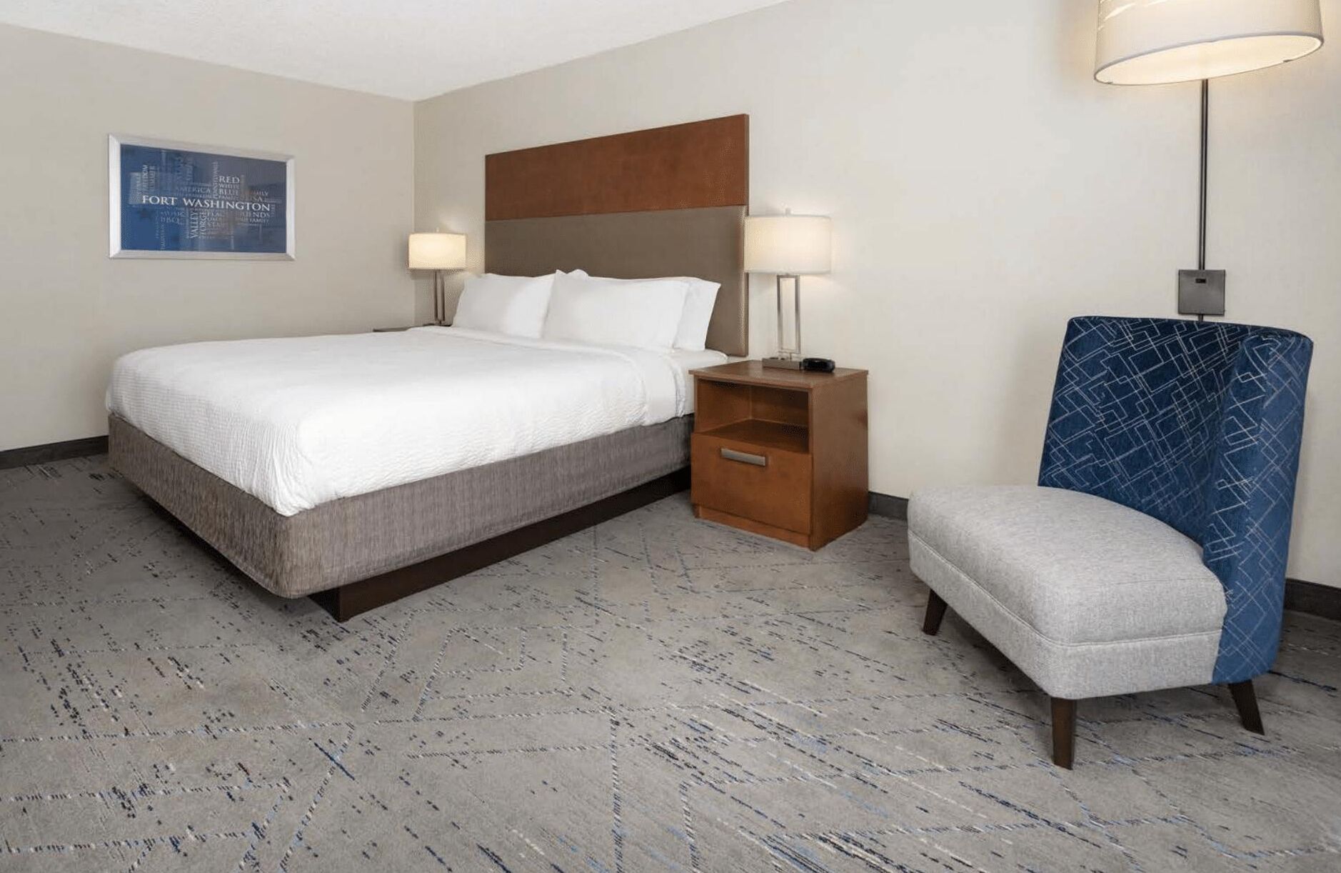 Une chambre d’hôtel avec un grand lit, une table de chevet avec une lampe, une chaise et une lampe fixée au mur.