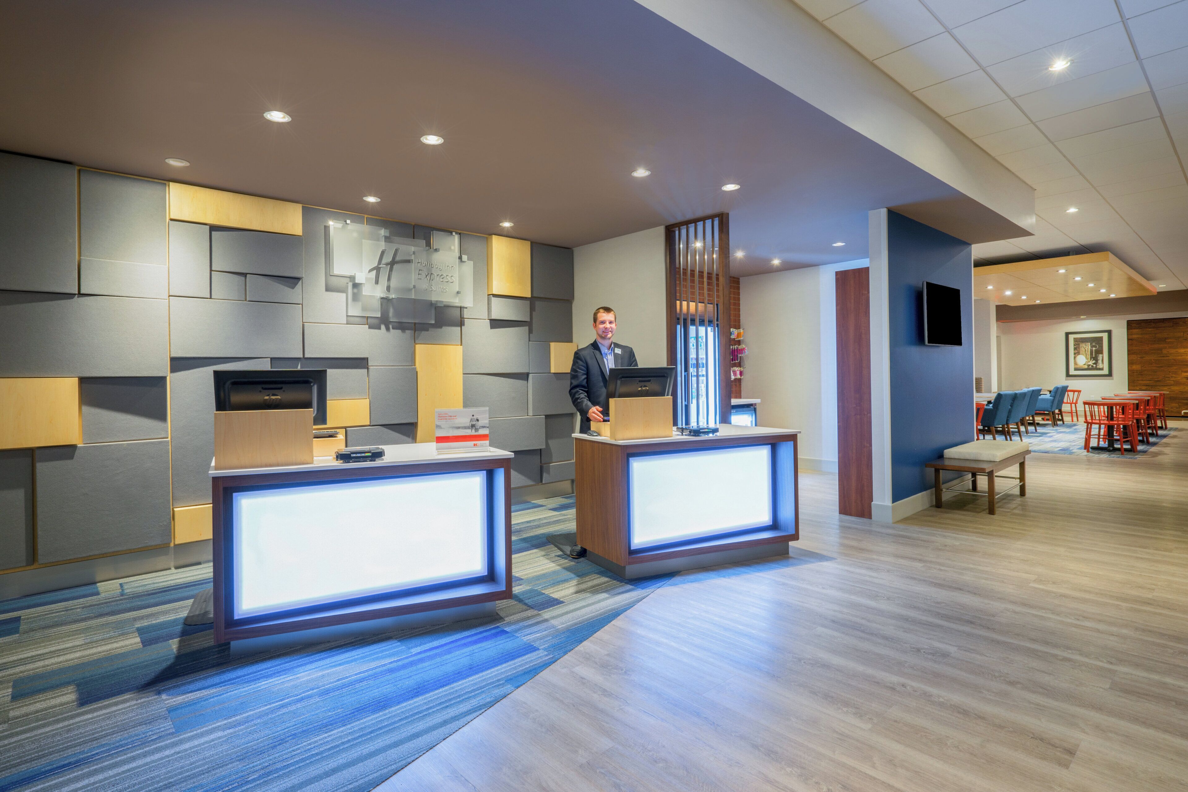 A modern hotel reception area with two reception desks, a seating area with chairs, and a television mounted on the wall.