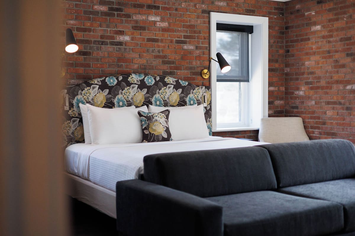 A bedroom with a brick wall, a bed with floral headboard, two bedside lamps, and a window.