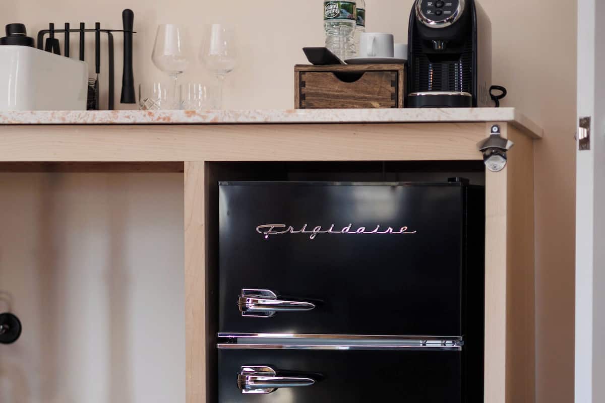 A compact black refrigerator with a drawer, a coffee maker, and two glasses on a wooden countertop.