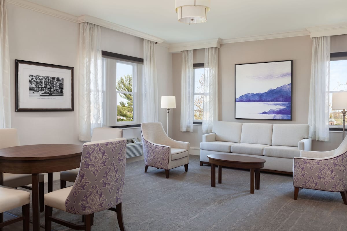 A modern living room with a round wooden table, two armchairs, a sofa, and a coffee table. There are large windows with sheer curtains, a framed picture on the wall, and a painting above the sofa.
