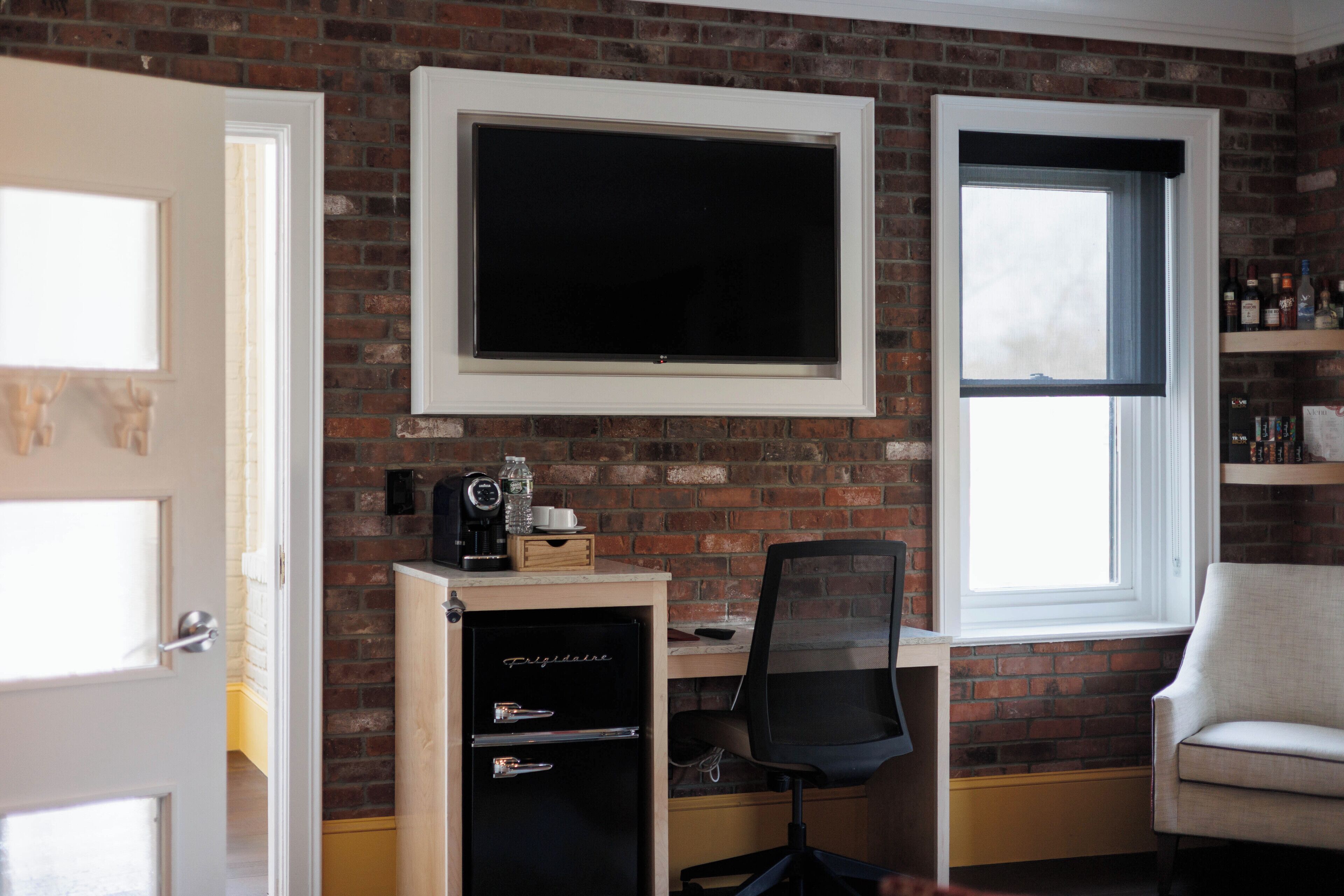 A room with a brick wall, a mounted TV, a desk with a chair, a small cabinet, and a window.