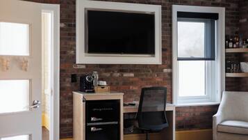 A room with a brick wall, a mounted TV, a desk with a chair, a small cabinet, and a window.