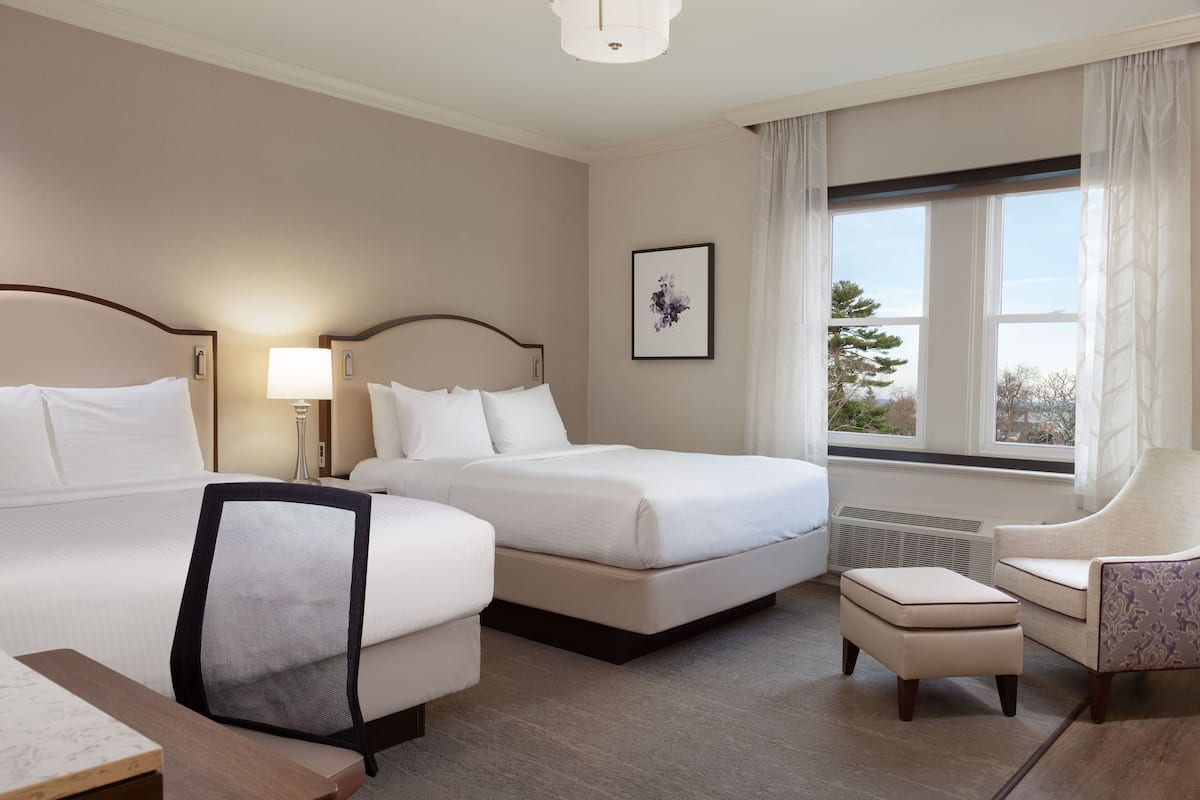 A hotel room with two beds, a desk, a chair, and a window with a view of trees.