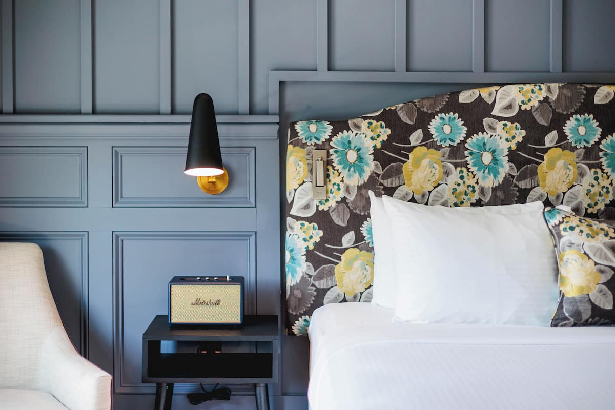 A hotel room with a bed, a nightstand with a radio, a chair, and a wall-mounted lamp.