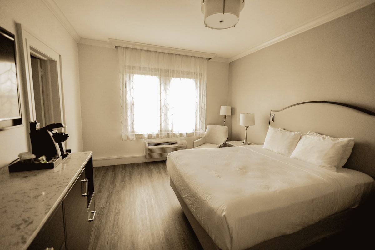 A hotel room with a large bed, a desk with a lamp, a chair, and a window with curtains.