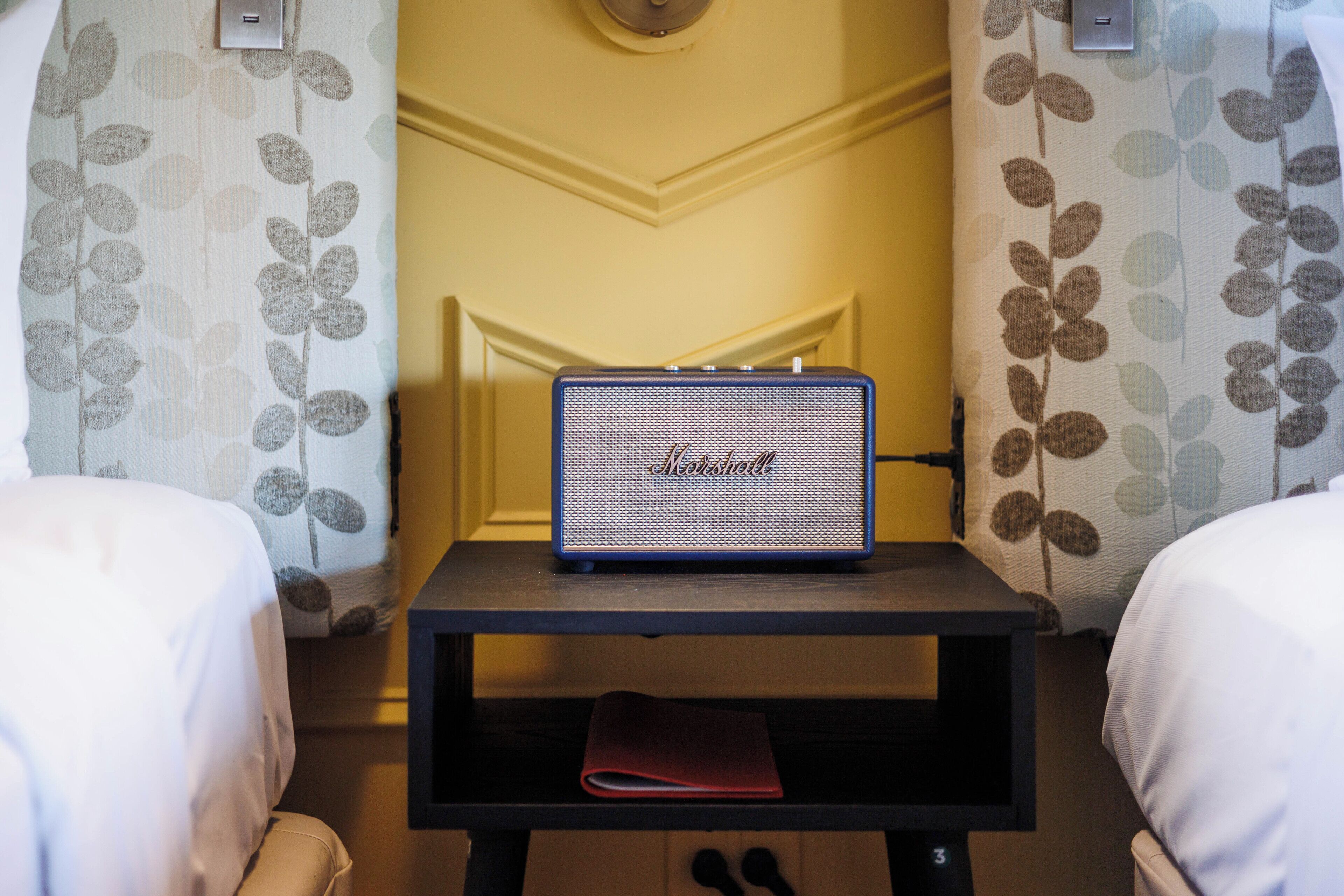 A hotel room with a bed, a small table with a speaker, and patterned curtains.