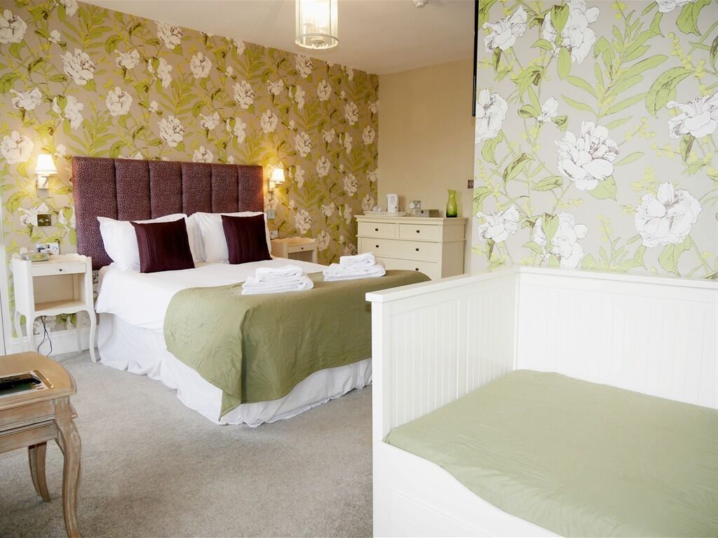 A hotel room with a large bed, a bedside table, a nightstand, a dresser, and floral wallpaper.
